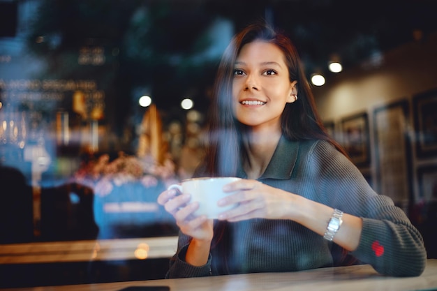 Mulher sorridente segurando uma xícara de café perto do rosto, sentada dentro de um