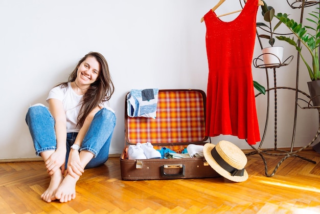 Mulher sorridente sentada no chão perto da valise com roupas embalando antes do espaço de cópia do conceito de viagem de viagem