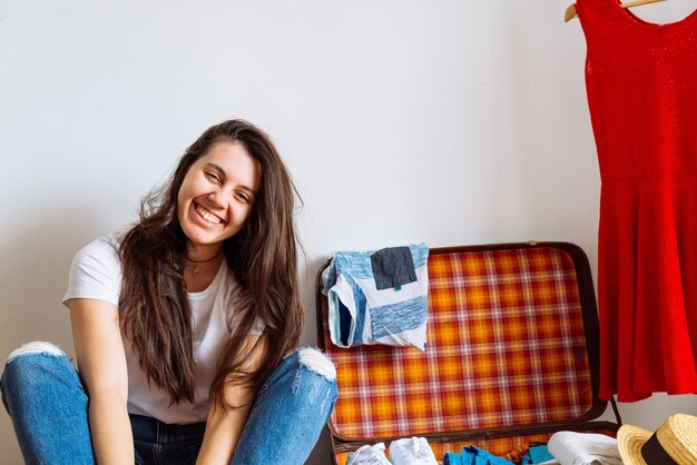 Mulher sorridente sentada no chão perto da valise com roupas embalando antes do espaço de cópia do conceito de viagem de viagem