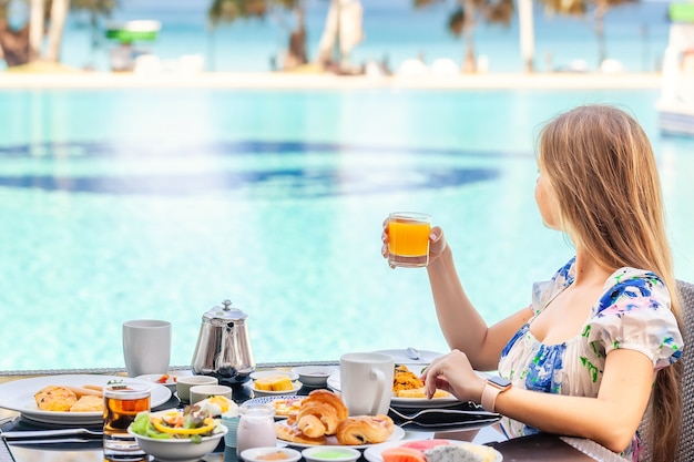 Mulher sorridente tomando café da manhã americano bebendo suco de laranja ao lado da piscina