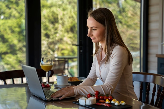 Mulher sorridente usa um laptop enquanto bebe suco de laranja enquanto está sentada em uma mesa durante o café da manhã