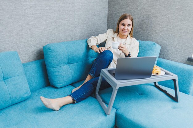 Mulher sorridente usando laptop para verificar notícias de e-mail on-line enquanto está sentado no sofá trabalhando no computador escrevendo blog ou assistindo webinar estudando em casa