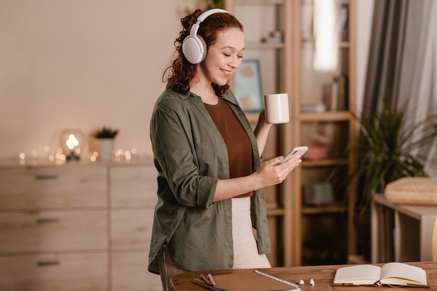 Mulher sorridente usando smartphone e fones de ouvido em casa enquanto toma um café