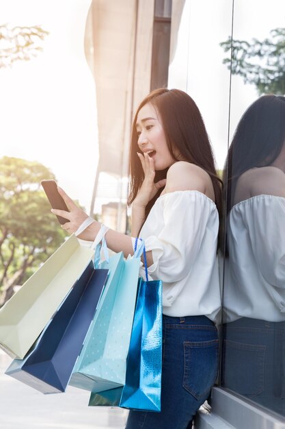 Mulher sorridente usando um telefone inteligente enquanto segura uma bolsa de compras de pé ao ar livre