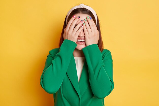 Mulher sorridente vestindo jaqueta verde posando isolada sobre fundo amarelo escondendo os olhos cobrindo o rosto com as palmas das mãos emoções positivas