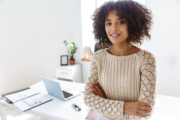 Mulher sorridente, vestindo roupas casuais, olhando para a câmera com os braços cruzados, em pé perto da mesa no escritório