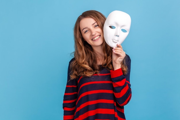 Mulher sorridente vestindo suéter listrado estilo casual segurando máscara branca mostrando sua expressão positiva fingindo ser outra pessoa Tiro de estúdio interior isolado em fundo azul