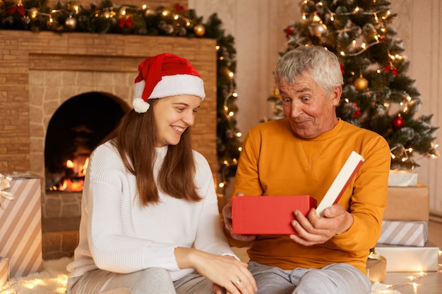 Mulher sorridente, vestindo uma blusa branca e chapéu de papai noel, sentada com o pai na sala de estar com as decorações de natal, comemorando a véspera de ano novo juntos.