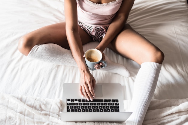Mulher sorrindo, bebendo um café e trabalhando com o computador na cama