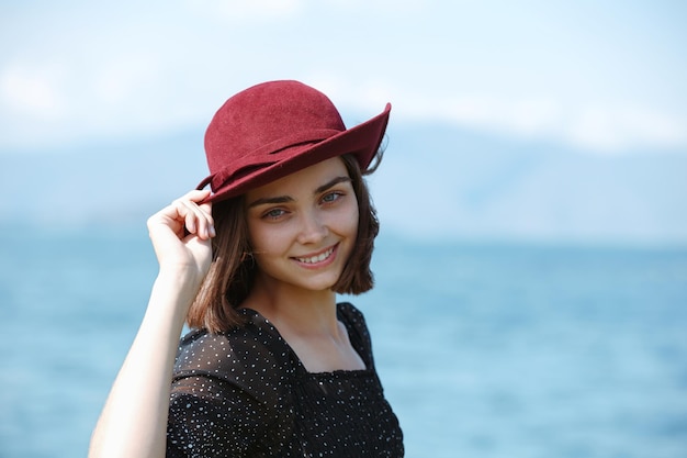 Mulher sorrindo e tocando o chapéu em pé contra o mar azul