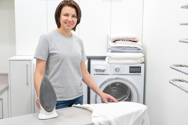 Mulher sorrindo em pé perto da tábua de passar roupa na lavanderia com máquina de lavar