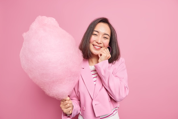 Mulher sorrindo gentilmente segurando o rosto segurando algodão doce no palito e vestindo roupas formais