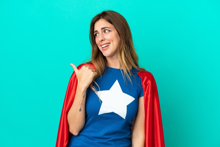 Foto mulher super-heroína caucasiana isolada em fundo azul apontando para o lado para apresentar um produto