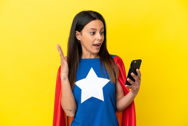 Mulher super-heroína isolada em fundo amarelo olhando para a câmera enquanto usa o celular com expressão de surpresa