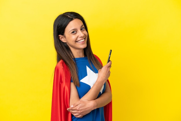 Mulher super-heroína isolada em um fundo amarelo segurando um telefone celular e com os braços cruzados