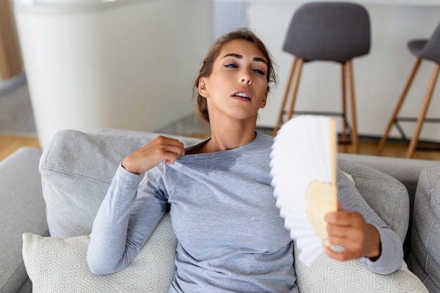 Mulher superaquecida sentada no sofá em casa sente-se quente acenando com ventilador de mão esfriando garota suada relaxe no sofá na sala de estar segure oscilação sofre de calor sem sistema de ar condicionado