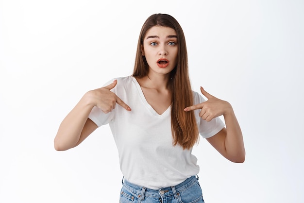 Foto mulher surpreendida, apontando para si mesma, levanta as sobrancelhas e ofegante, mostrando o logotipo de autopromoção demonstrando banner sobre fundo branco