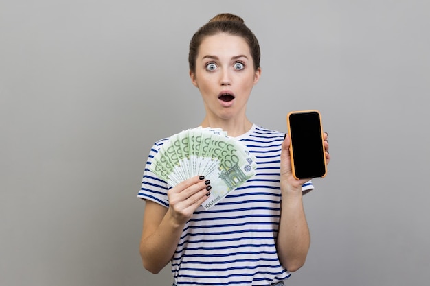 Mulher surpreendida chocada segurando fã de notas de euro e telefone inteligente com tela preta em branco