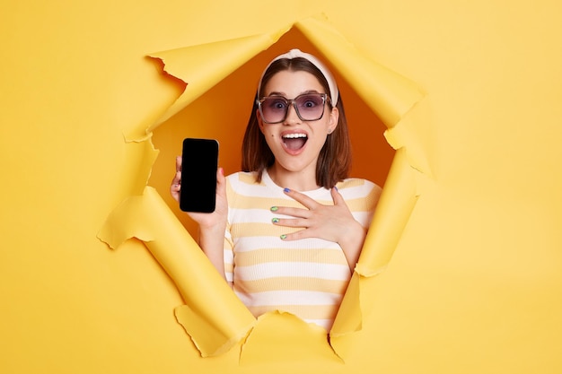 Mulher surpreendida e muito feliz fica no buraco de papel rasgado mostrando telefone inteligente com display vazio com área de propaganda olhando através do avanço do fundo amarelo