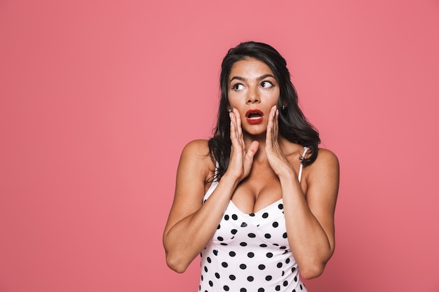 Foto mulher surpreendida em trajes de banho posando isolada sobre uma parede rosa, olhando de lado com a boca aberta.