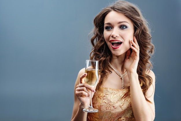 Foto mulher surpreendida em vestido dourado e colar de pérolas com taça de champanhe e tocar-se rosto para a mão