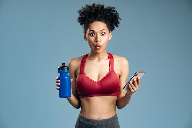 Foto mulher surpreendida segurando a garrafa com água e smartphone, enquanto olha para a câmera com a boca aberta e expressão chocada. foto de estúdio interna isolada em fundo azul