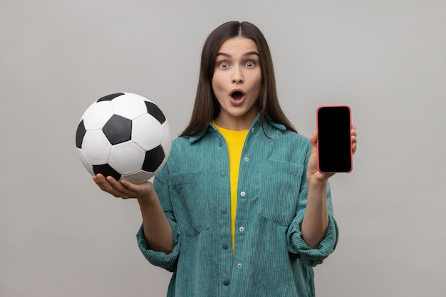 Mulher surpreendida segurando e mostrando smartphone em branco e bola de futebol apostando no futebol