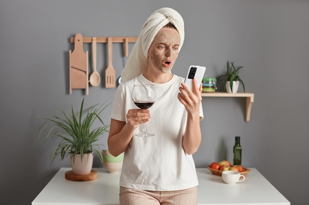 Mulher surpresa chocada com máscara cosmética em toalha branca enrolada na cabeça bebendo vinho na cozinha segurando o telefone inteligente na mão olhando para a tela com a boca aberta lendo as últimas notícias