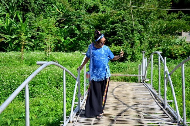 Mulher tailandesa usa telefone inteligente para tirar foto da vila de Baan Natong na ponte de bambu de madeira em Phare Tailândia