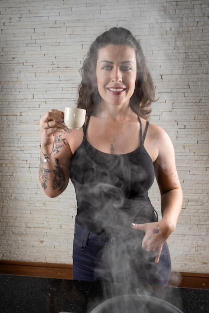 Mulher tatuada brasileira sorridente segurando uma xícara de café atrás da fumaça saindo do pote