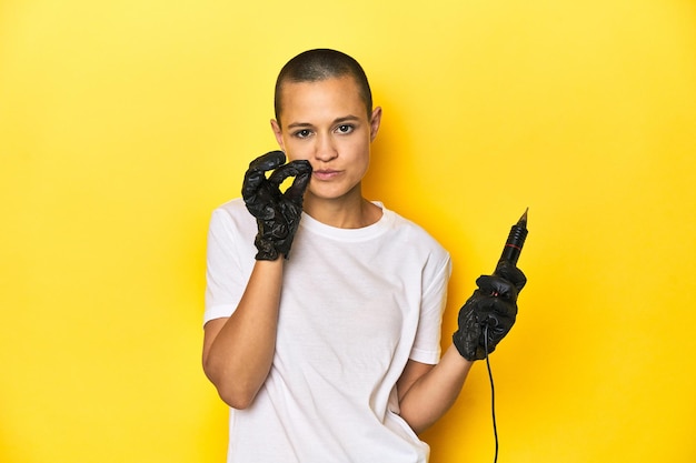 Mulher tatuadora no estúdio, cabeça rapada, fundo amarelo, dedos nos lábios, guardando um segredo.