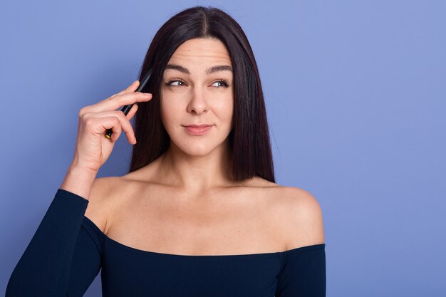 Mulher tem expressão pensativa séria, segurando a caneta na mão e os dedos perto da têmpora, usando um vestido estiloso com ombros nus