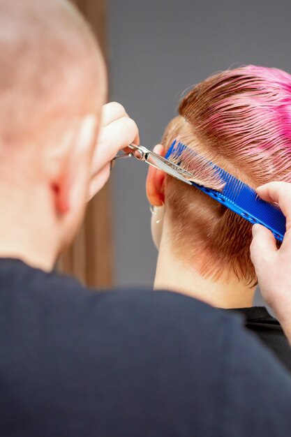 Mulher tendo um novo corte de cabelo. Cabeleireiro masculino cortando cabelo curto rosa com tesoura em um salão de cabeleireiro.