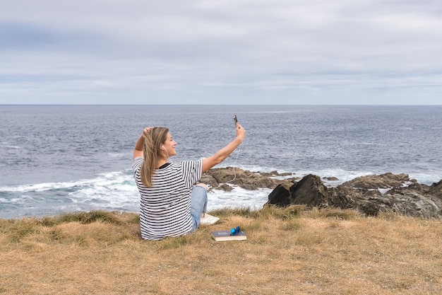 Mulher tirando um autorretrato com um telefone celular, sentado em frente ao mar.