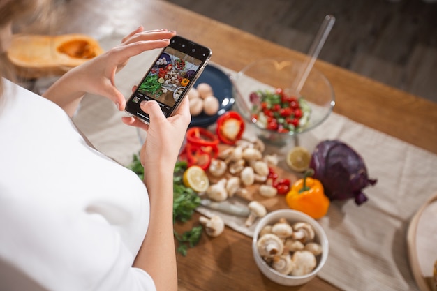 Foto mulher tirar uma foto de diferentes belos legumes e verduras com um telefone móvel em sua cozinha. fotografia de comida de smartphone. alimentação saudável, comida vegetariana, dieta. para perda de peso e desintoxicação.