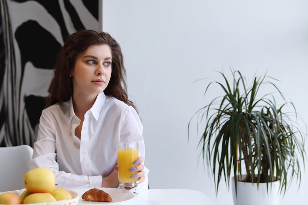 Mulher toma café da manhã em casa com suco de laranja e croissant