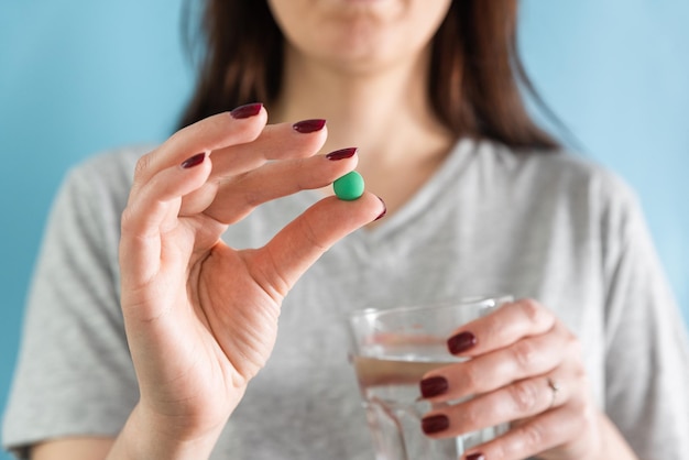 Mulher tomando pílula verde para dor de cabeça ou analgésico com copo de água sobre fundo azul tomando remédio