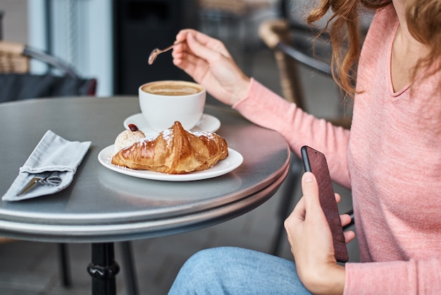 Mulher tomar café da manhã em caffe e usando o smartphone. Menina conversando e usando a internet com telefone durante o coffee-break com um croissant