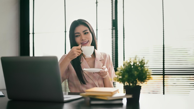 Mulher trabalhadora agradável bebendo café e lendo notícias on-line ou e-mail comercial em seu laptop