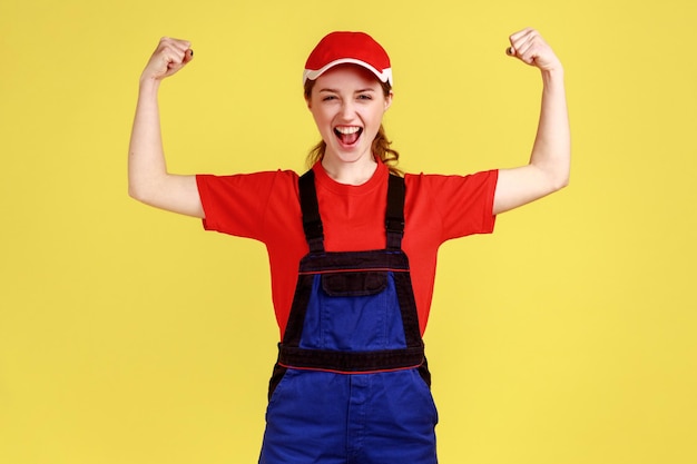 Mulher trabalhadora animada em pé com os braços levantados mostrando seu bíceps gritando alegremente olhando para a câmera vestindo macacão e boné vermelho Estúdio interior isolado em fundo amarelo