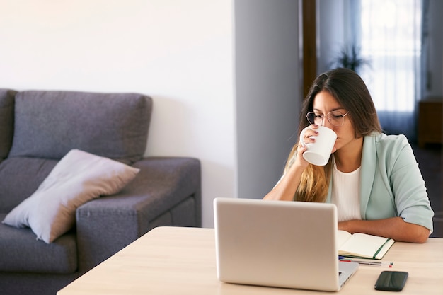 Mulher trabalhando em casa com o laptop e bebendo café.