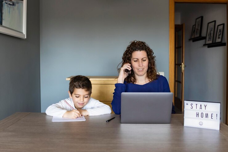 Foto mulher trabalhando em laptop enquanto está sentada ao lado do filho estudando na mesa