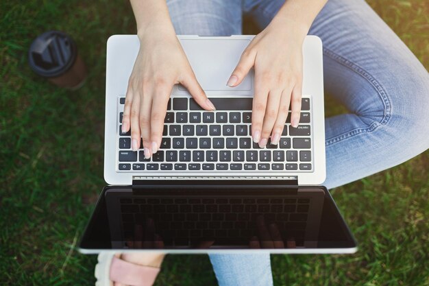 Mulher trabalhando em um laptop com tela em branco, sentada na grama verde ao ar livre, vista superior