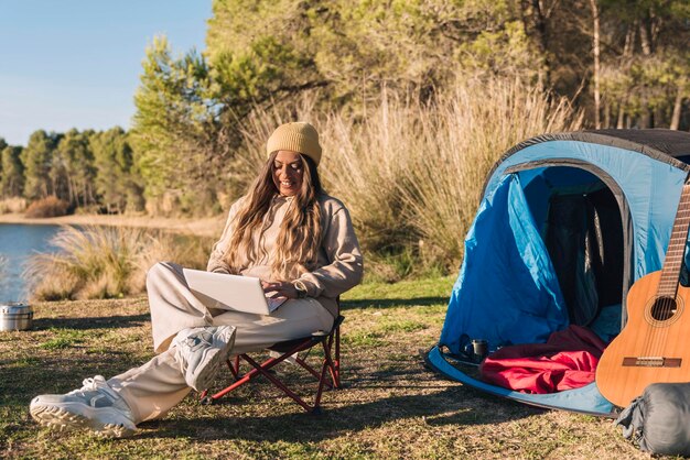 Mulher trabalhando no laptop perto da barraca de acampamento ao ar livre cercada pela natureza