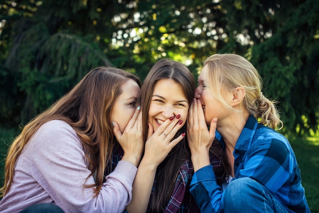 Mulher três atrativa nova que compartilha dos segredos que sentam-se na grama verde no parque. Namoradas alegres fofocar e sussurrar ao ar livre