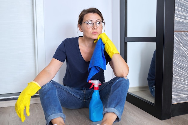 Mulher triste e cansada em luvas com detergente e pano, sentada no chão da sala terminou de limpar a casa. Higiene, pureza, casa, conceito de limpeza