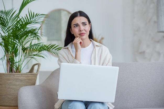 Mulher triste e chateada em desespero lendo más notícias da morena de laptop em óculos sentada no sofá em