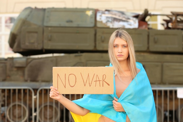 Mulher triste envolta em bandeira ucraniana segurando cartaz com palavras sem guerra perto de tanque quebrado na rua da cidade