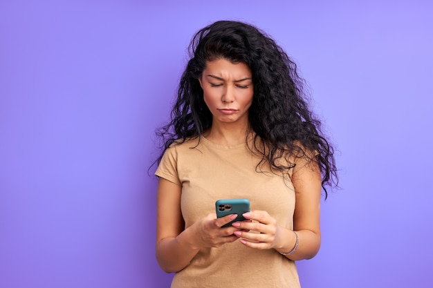 Foto mulher triste está preocupada com alguma pergunta, olhando para o telefone celular, tendo uma expressão facial chateada. parede roxa isolada