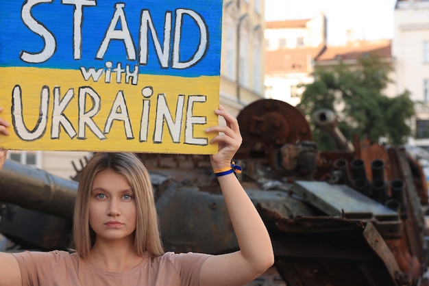 Foto mulher triste segurando cartaz nas cores da bandeira nacional com palavras stand with ukraine perto de tanque quebrado na rua da cidade